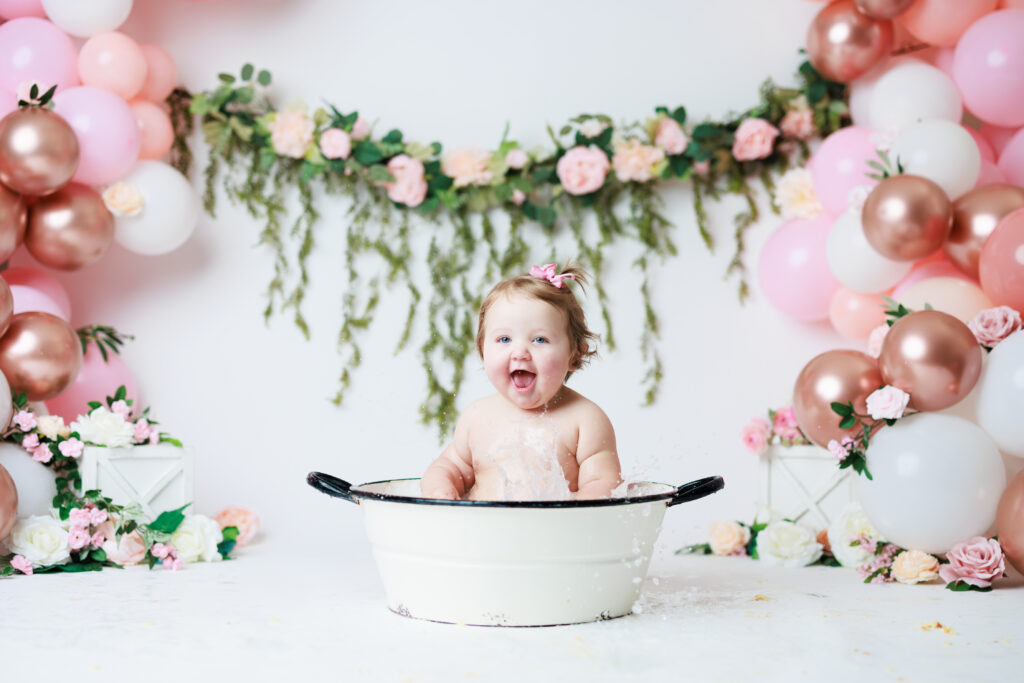 baby splashing in white bathtub with balloons and floral garland as backdrop
