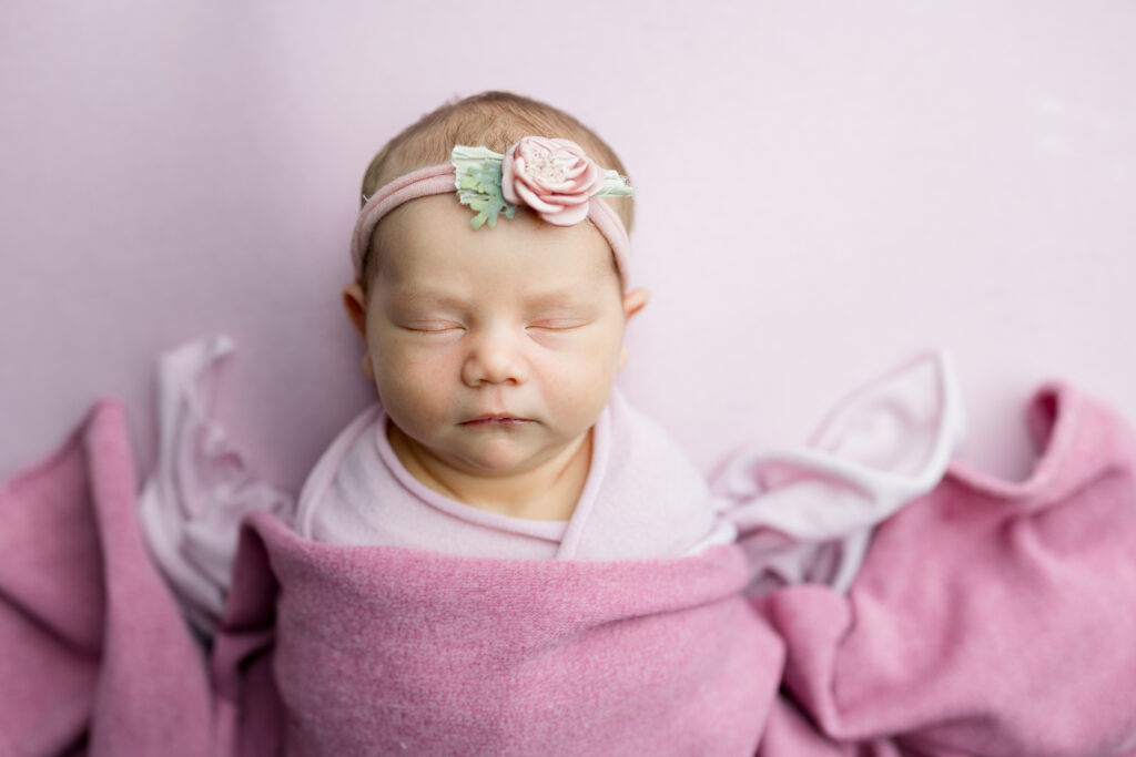 baby wrapped in pink swaddle on pink backdrop sleeping with floral headband
