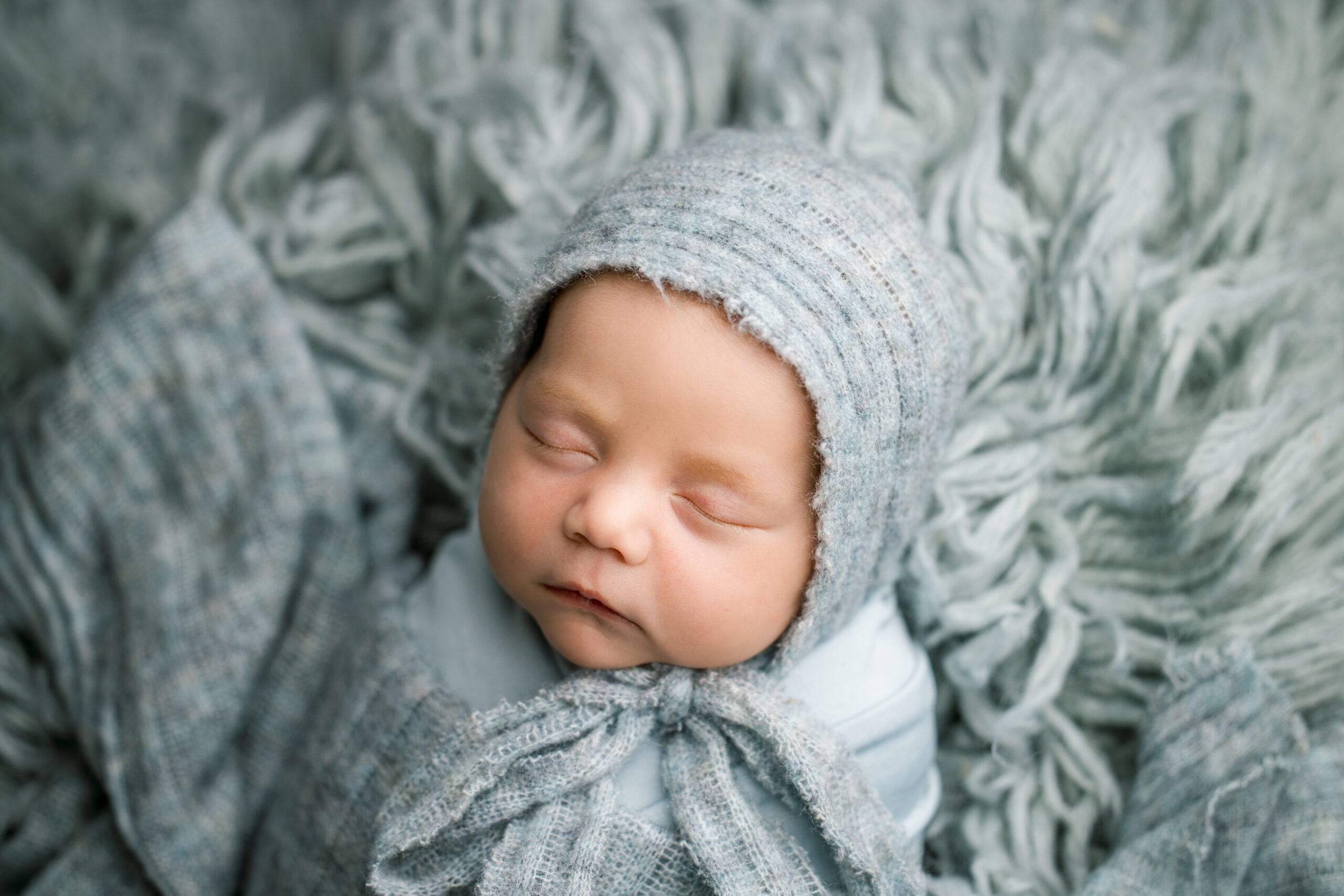 baby lying on blue fluffy rug swaddled in a blue wrap weating a blue bonnet