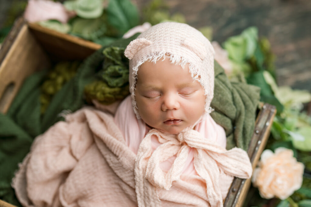 baby posed in a wood box surrounded by peach and pink flowers wearing a teddy bonnet 
