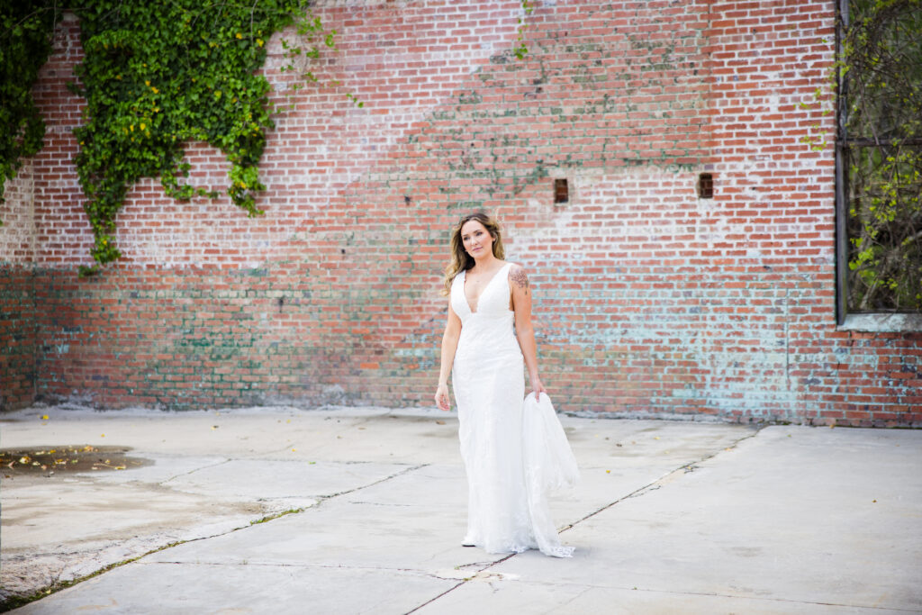 bride standing in white dress holding the back standing in front of a brick wall with greenery 