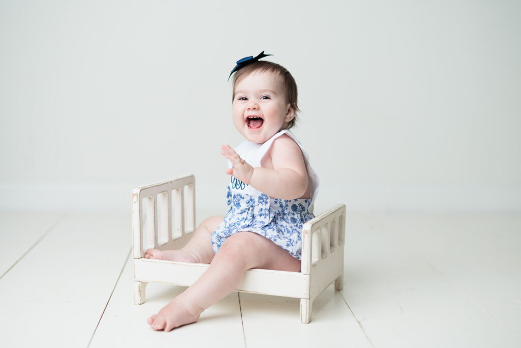 baby sitting in blue dress in a white bed prop smiling 
