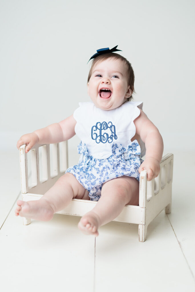 baby smiling in blue dress with scallop sleeve sitting on white bed prop