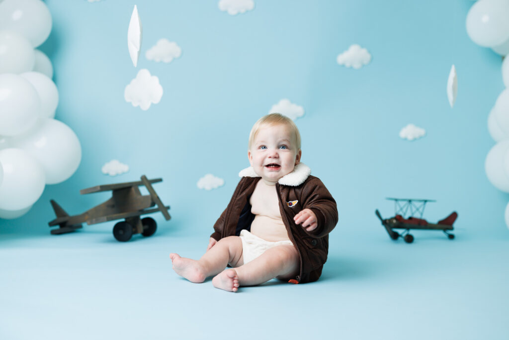 boy in bomber jacket with vintage airplanes in the background for a first birthday cake smash