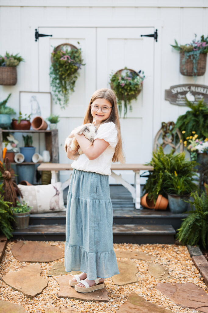 girl standing holding bunny in front of potting shed with flowers for spring 