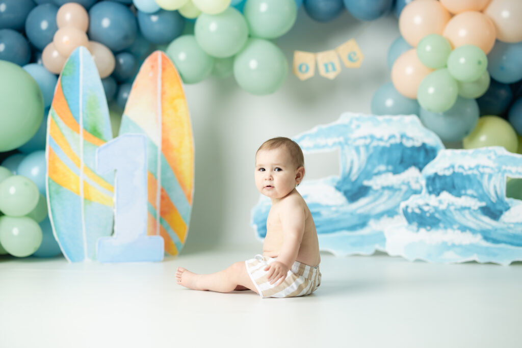 boy sitting in front of a big wave decor with surf boards for a first birthday session 