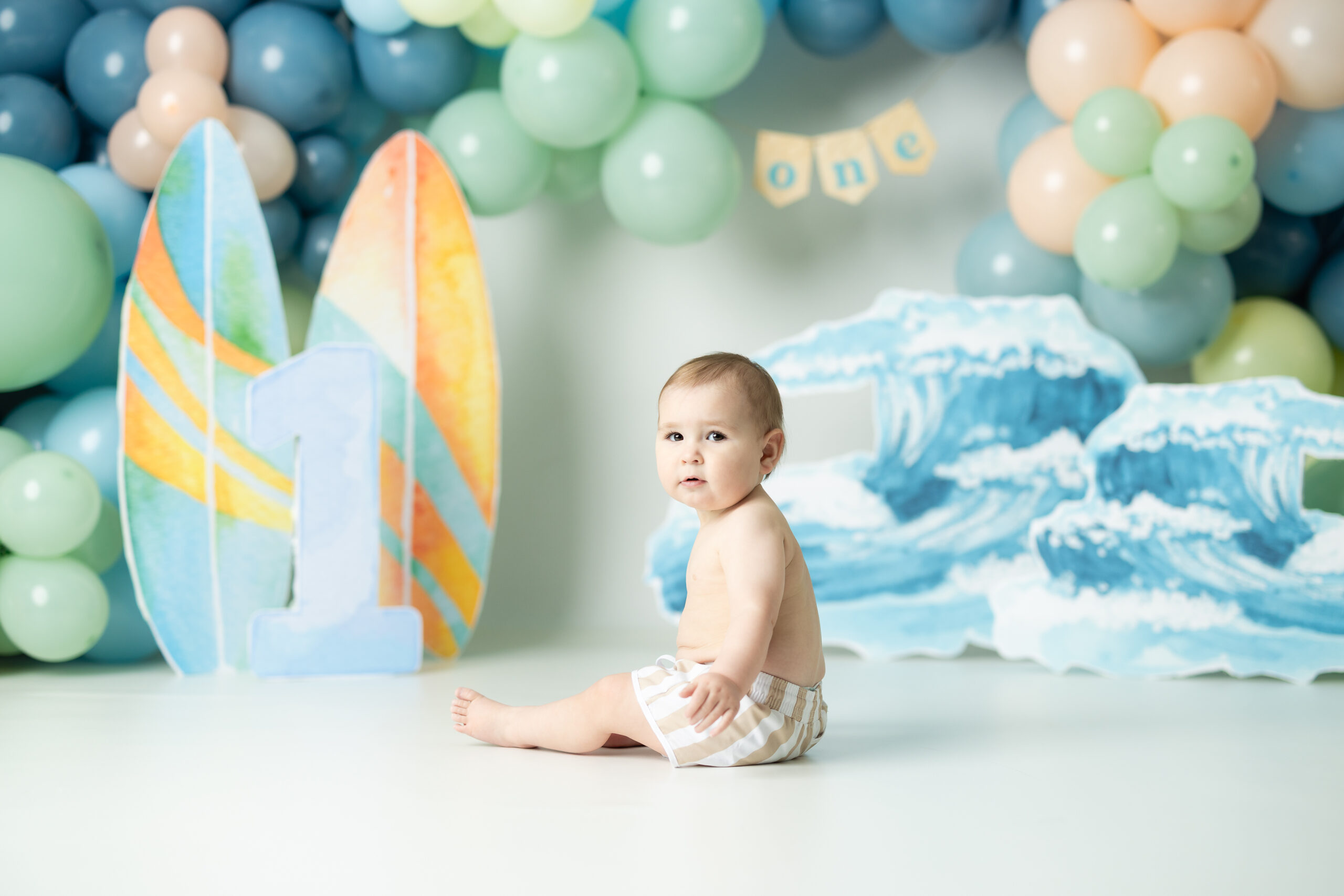 little boy sitting in swim trunks in front of a backdrop that is the big one, surf boards with waves