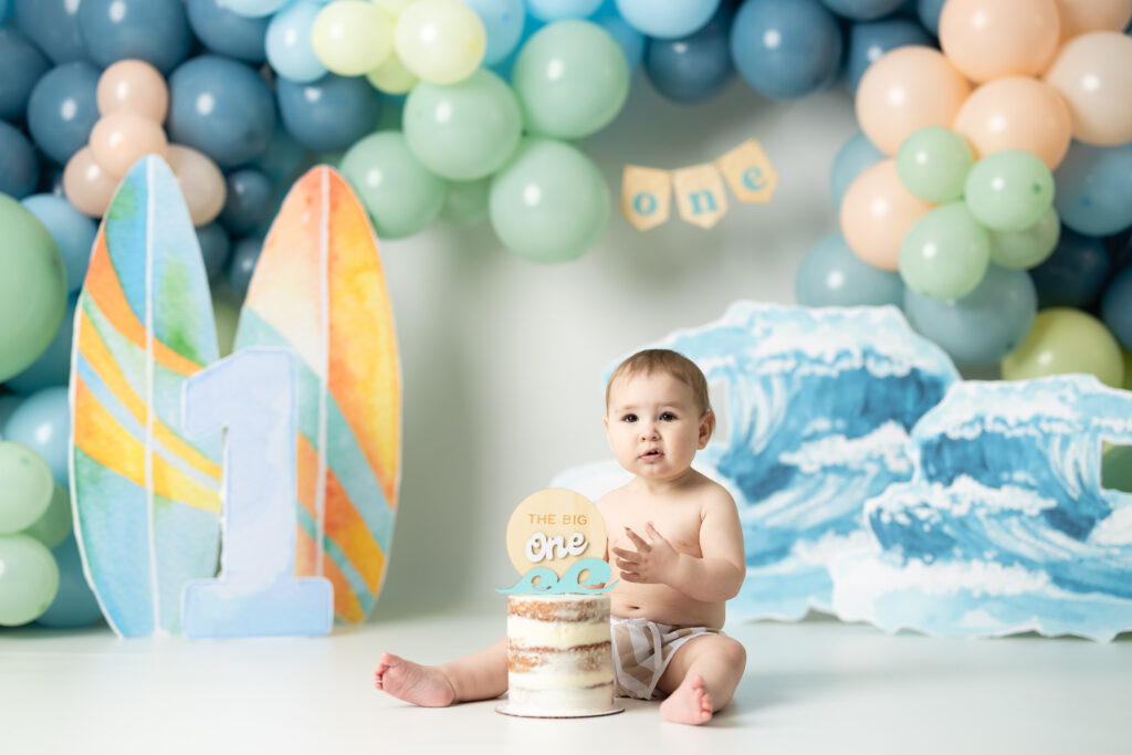 little boy sitting in swim trunks in front of a backdrop that is the big one, surf themed 