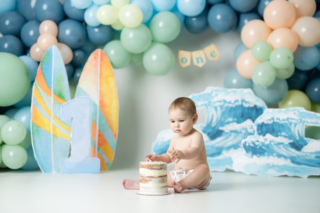 little boy sitting in swim trunks in front of a backdrop that is the big one, surf themed eating cake