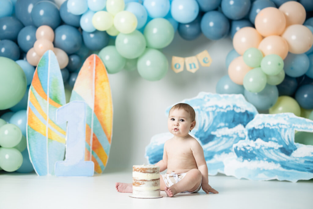 little boy sitting in swim trunks in front of a backdrop that is the big one, surf themed 