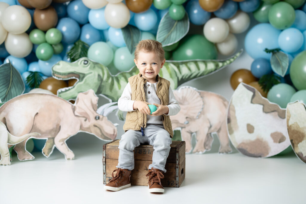 little boy sitting on brown box holding a small dinosaur egg smiling 