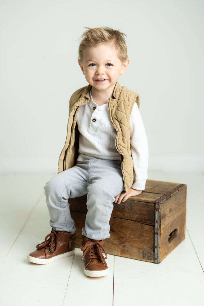 little boy wearing a vest sitting on a brown box smiling 