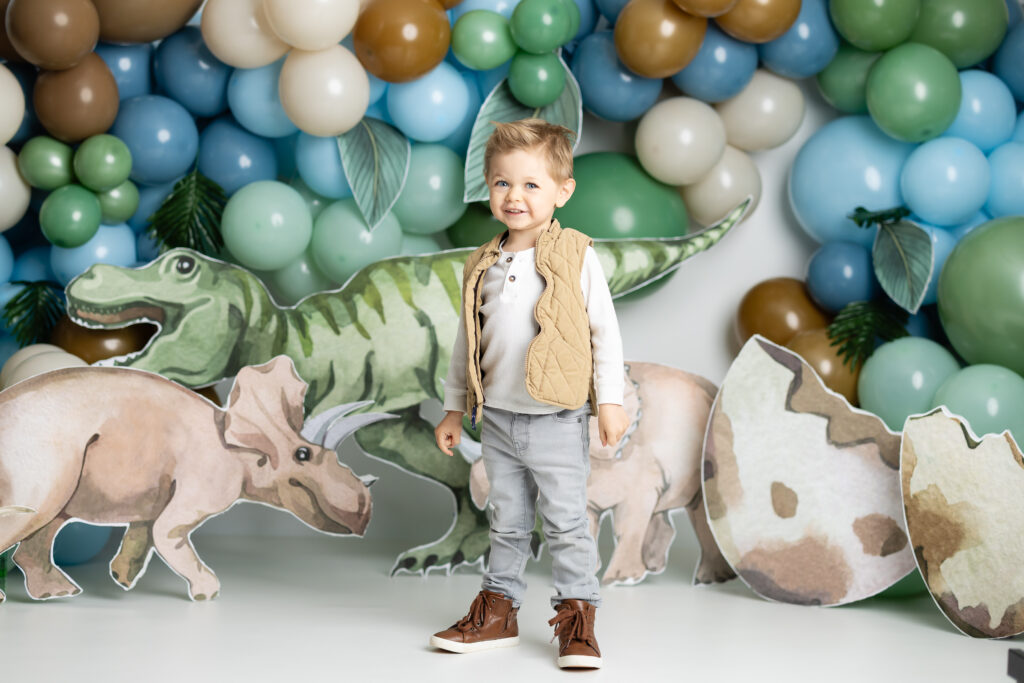 picture of almost 3 year old taking birthday pictures standing in front of dinosaur backdrop with balloon arch 