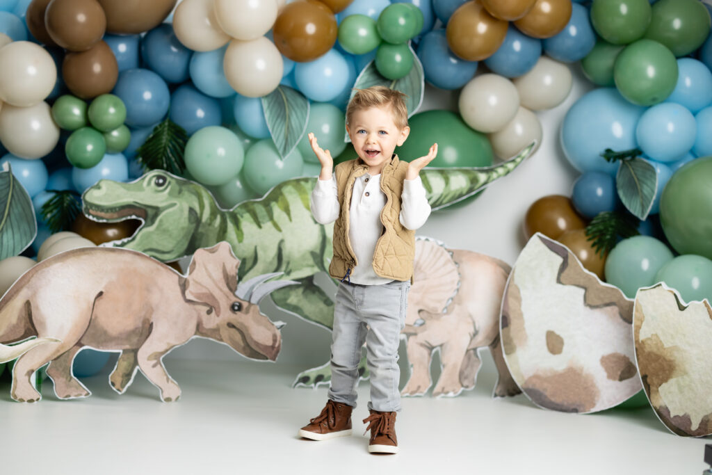 boy standing and smiling in front of a dinosaur backdrop with balloons 