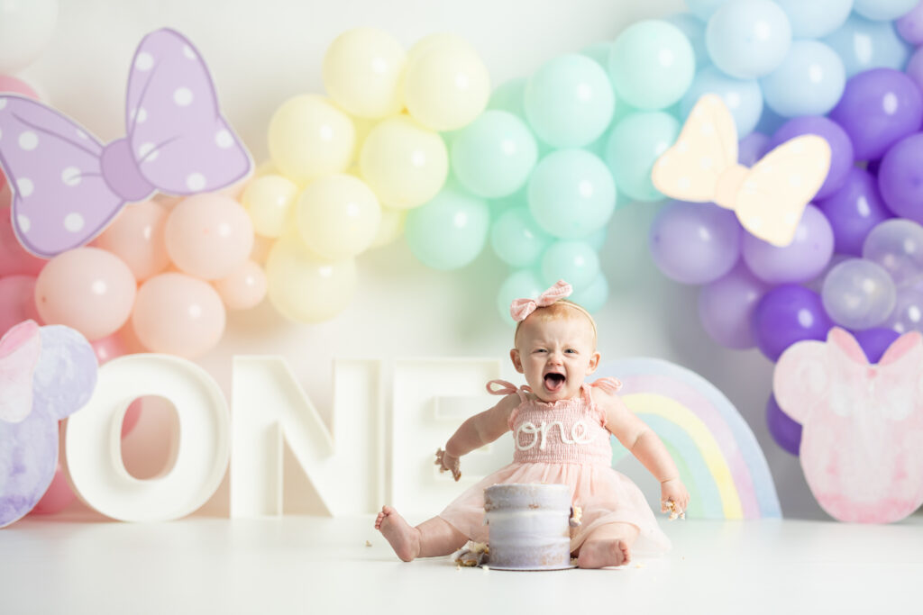 baby eating cake in front of rainbow colored balloon arch 