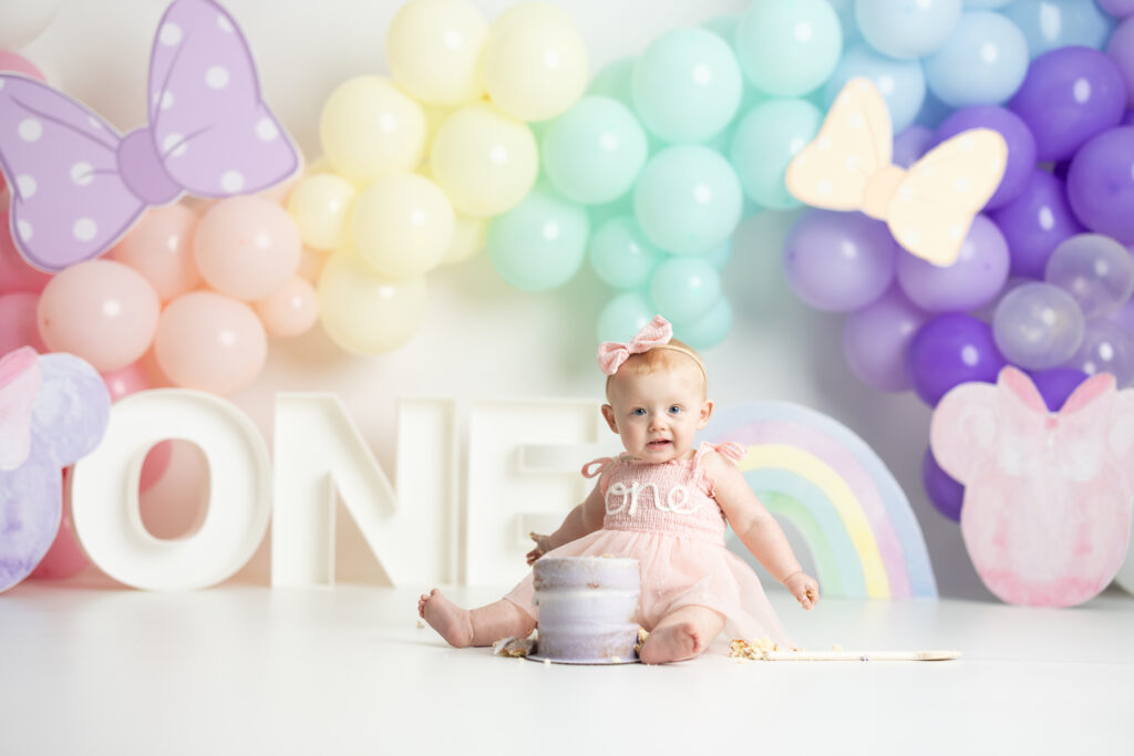 baby eating cake in front of rainbow colored balloon arch 