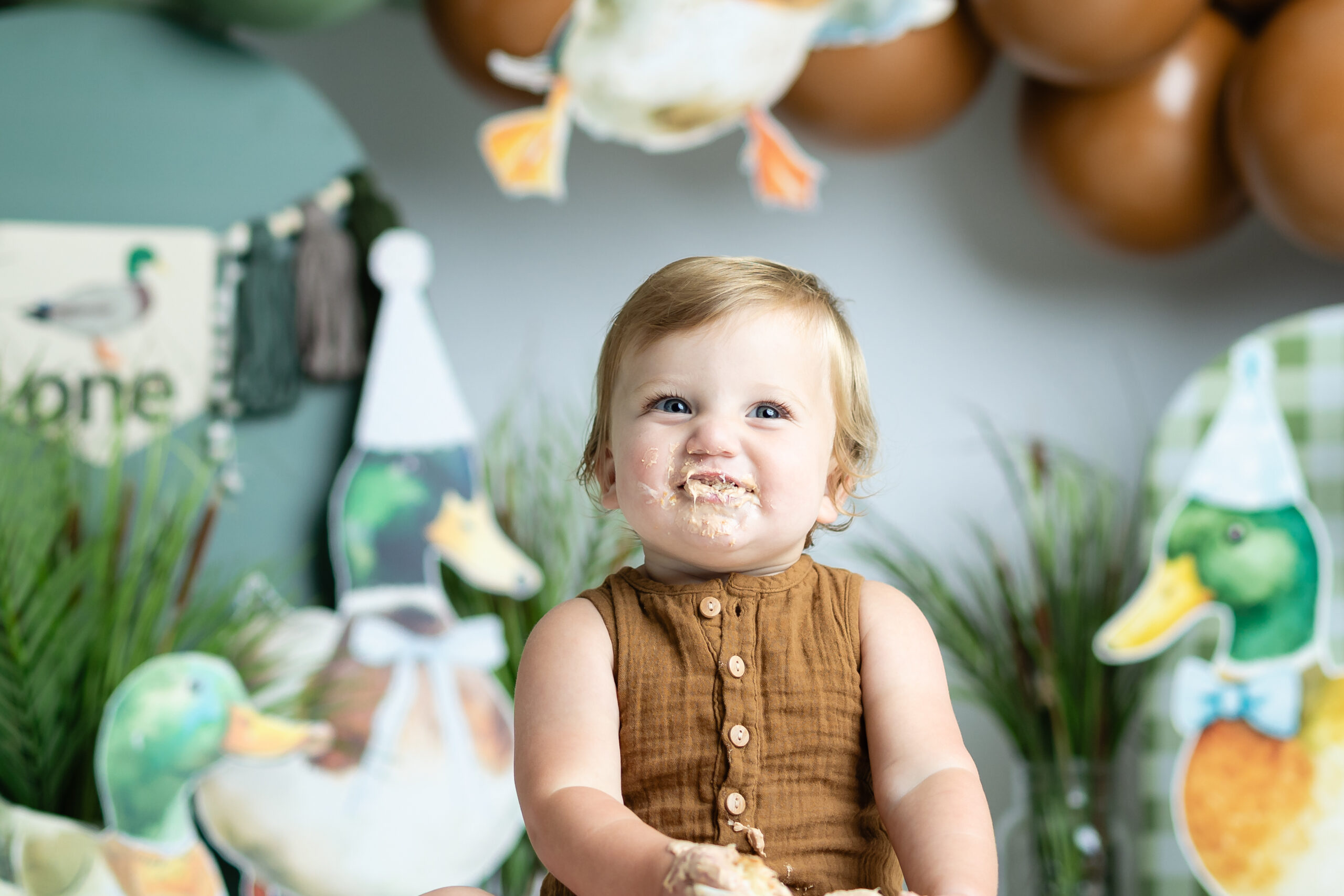 bit celebrating first birthday by doing a cake smash photography session sitting in front of a backdrop filled with balloons and mallard ducks and grass