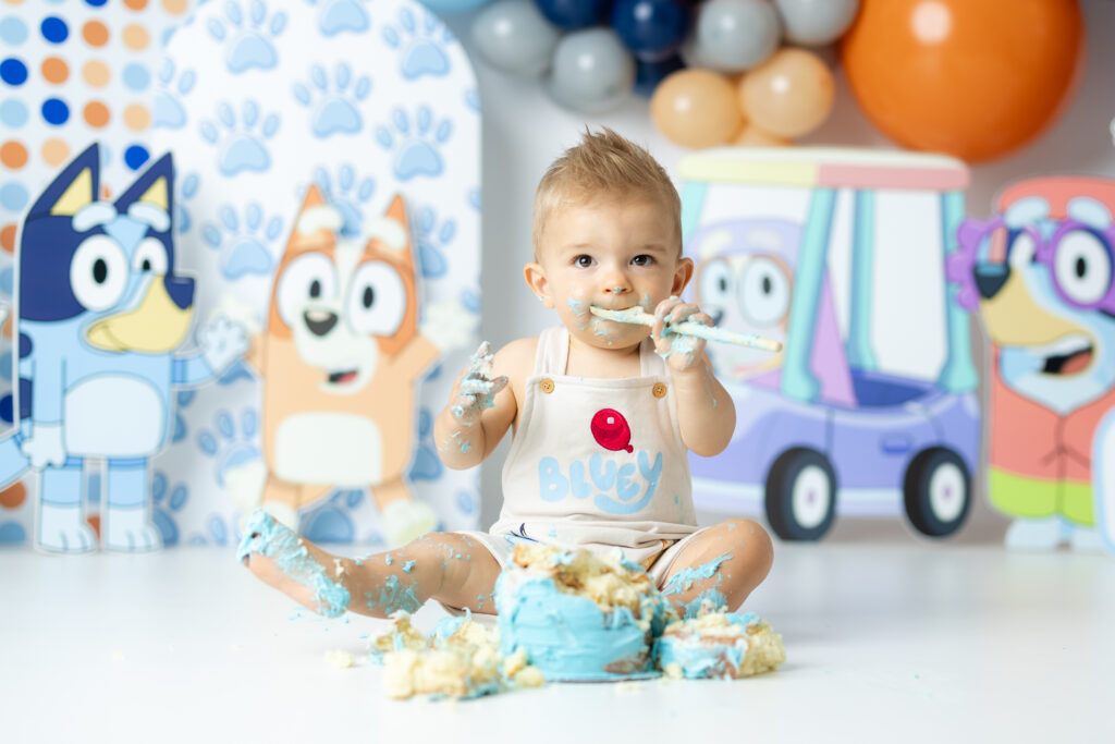 One-year-old boy with brown eyes and spiked hair smiling in front of Bluey cake smash backdrop