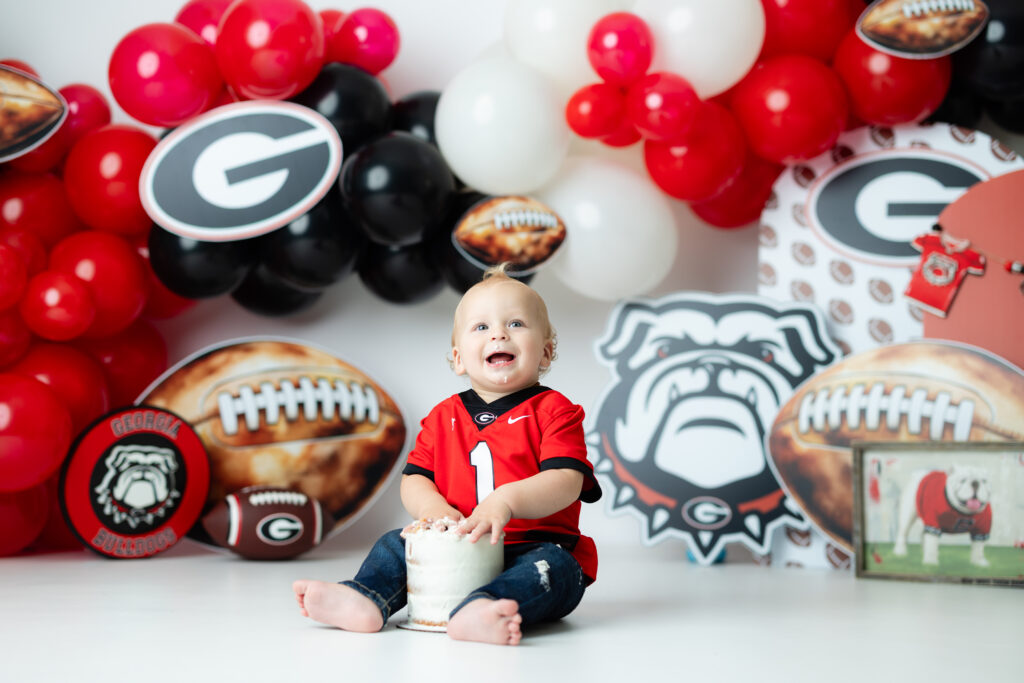 Smiling blonde baby with mohawk and curls reaching for his first birthday cake during Georgia Bulldogs cake smash session

