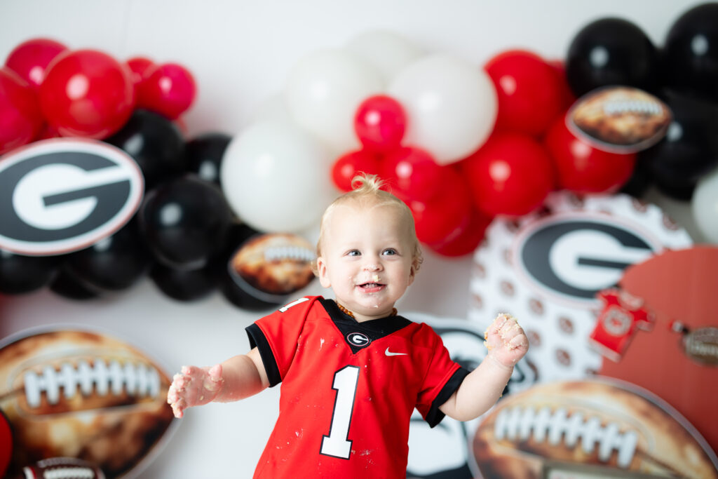One-year-old boy in a Georgia Bulldogs jersey with the number one sitting in front of a red and black UGA-themed cake smash set