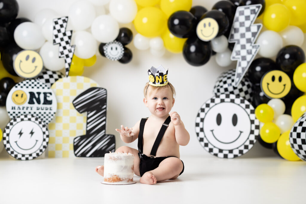 One year old boy smiling in front of “One Happy Dude” cake smash set with balloons.