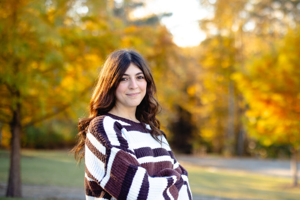 Senior in striped sweater posing on gravel path with autumn trees.