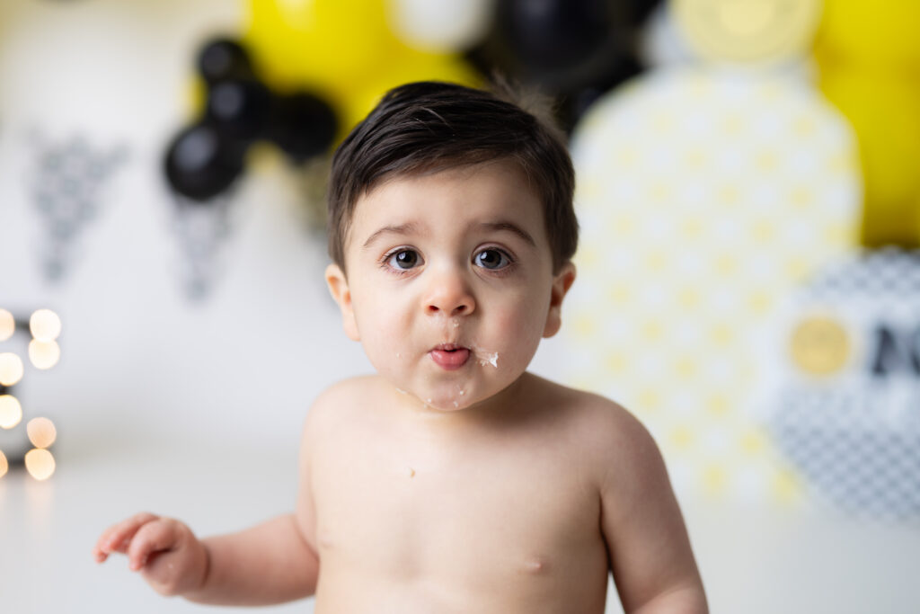 Baby boy during One Happy Dude Cake Smash Canton GA session with black and yellow smiley backdrop and frosting on his lips.