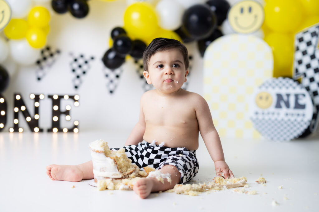 One Happy Dude Cake Smash Canton GA baby boy sitting on white studio floor with smashed cake, black and yellow balloons, and checkered shorts.