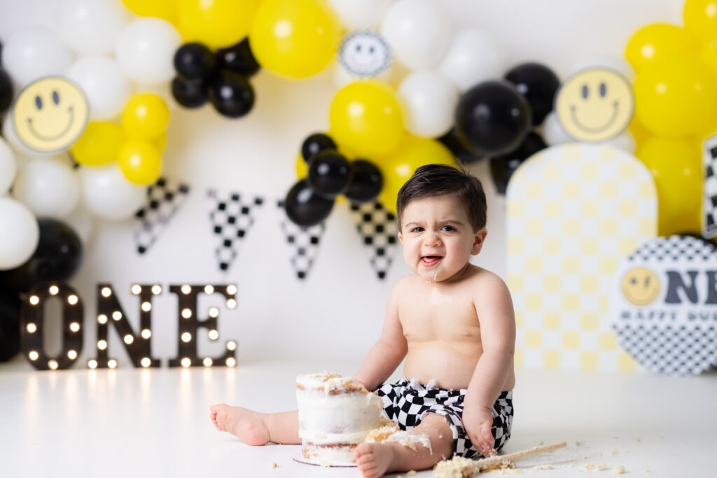 Baby boy reacting during One Happy Dude Cake Smash Canton GA session with black, yellow, and smiley face balloon garland in Canton GA studio.