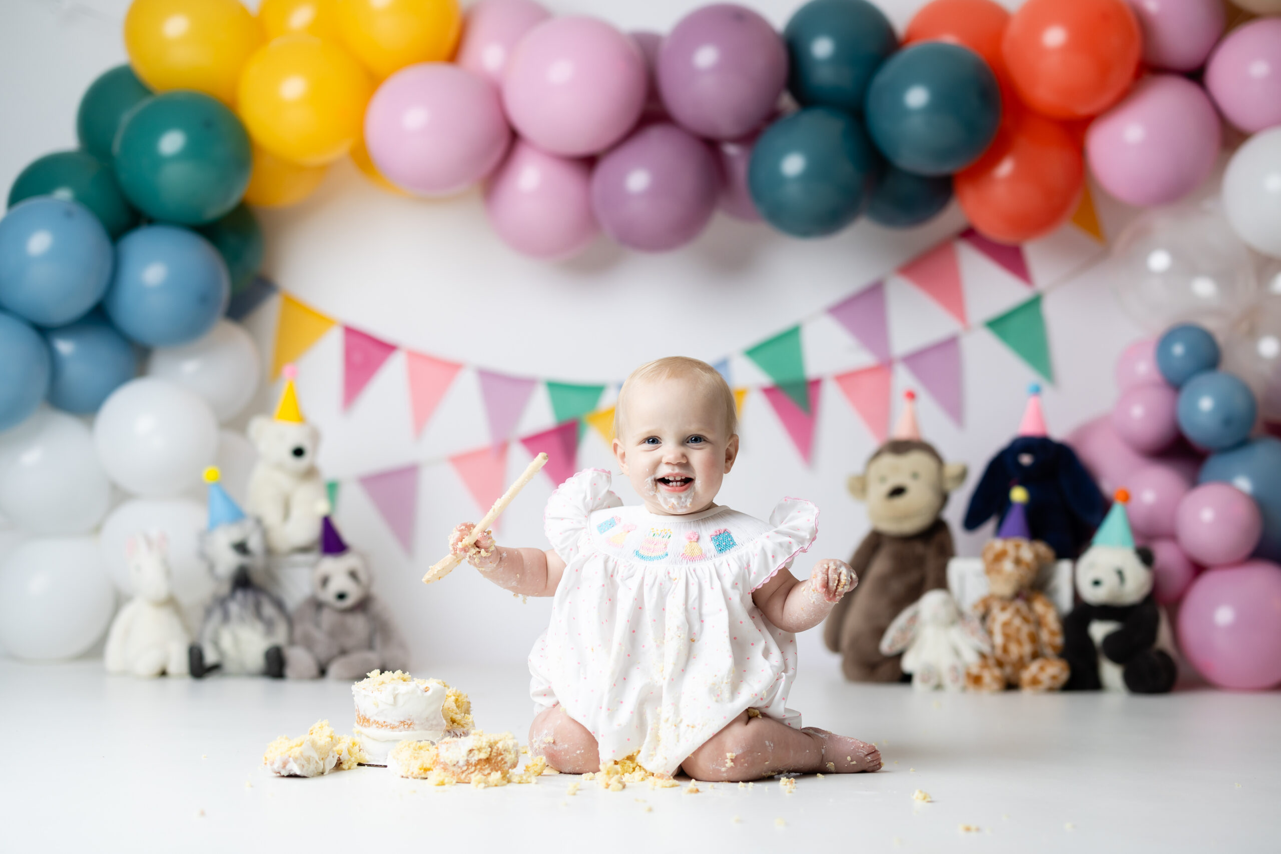 One-year-old girl sitting in a wooden bowl with stuffed party animals and colorful balloons